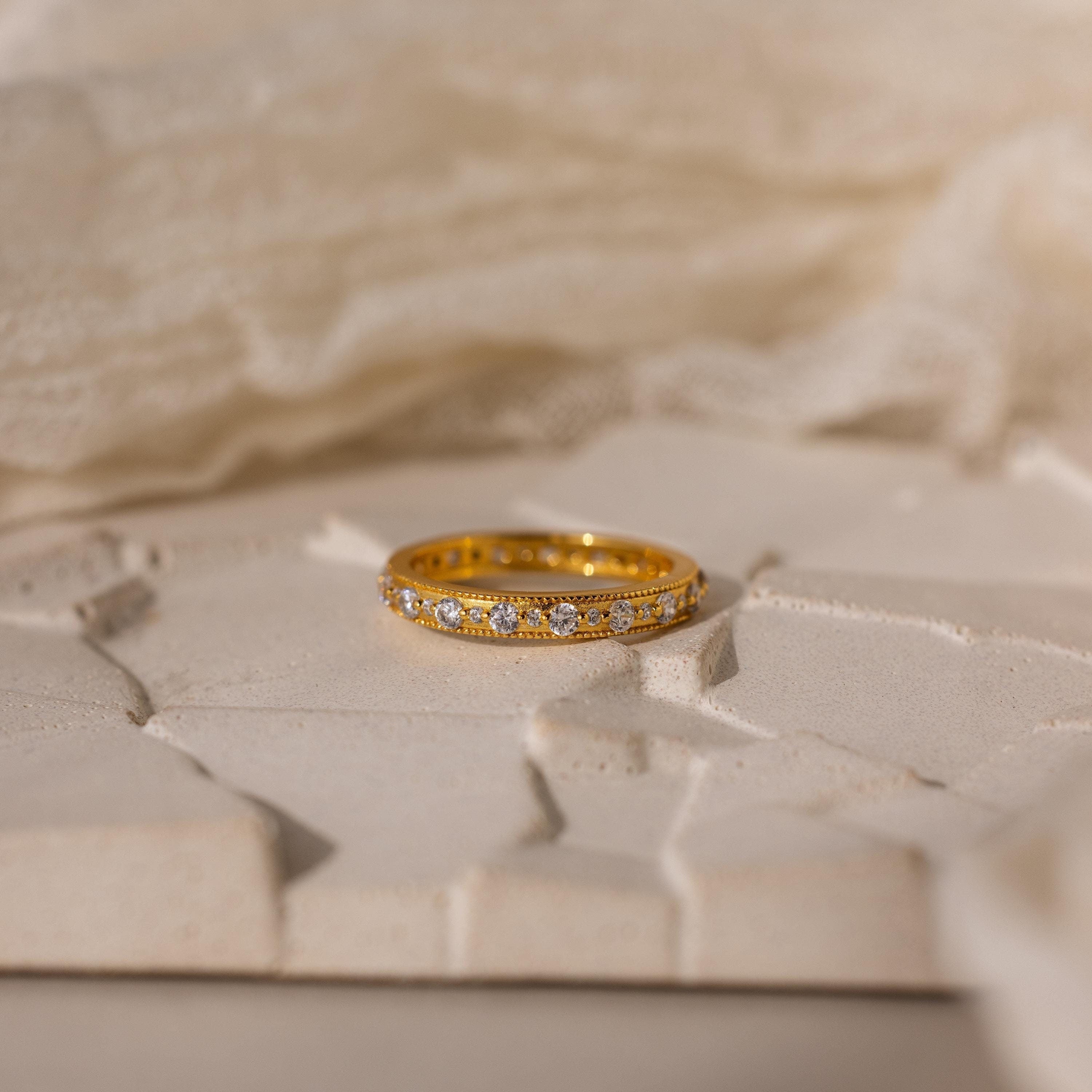 The Diamond Eternity Ring, a stackable gold band adorned with small diamonds, is displayed on a textured cream surface with a soft fabric background.
