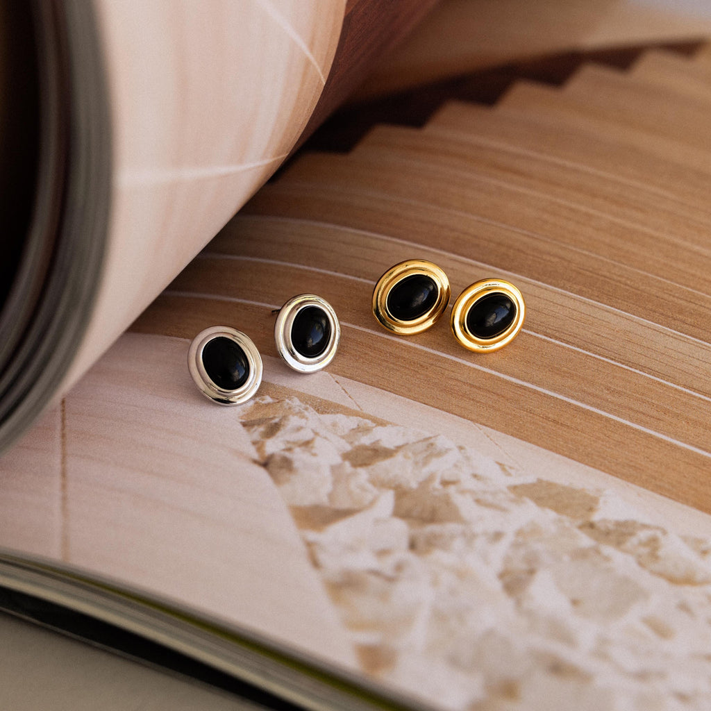 A pair of Vintage Oval Black Studs are displayed on an open book with abstract pages, showcasing their retro statement stud earring style.