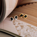 A pair of Vintage Oval Black Studs are displayed on an open book with abstract pages, showcasing their retro statement stud earring style.