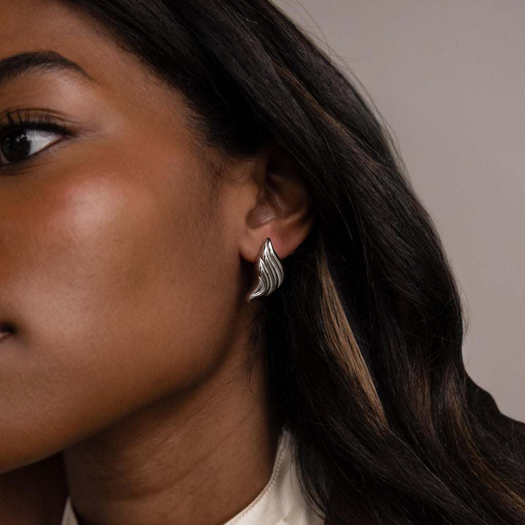 Close-up of a woman wearing Bold Wing Studs, featuring a shiny, wave-shaped design, on her left ear.