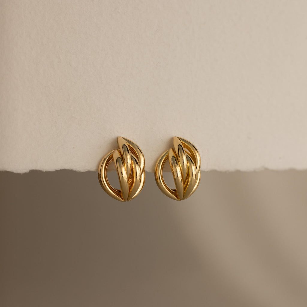 The Bold Multi Knot Studs, gold knot-shaped earrings with modern elegance and a bold multi-knot design, are displayed on a textured off-white surface against a soft, neutral background.
