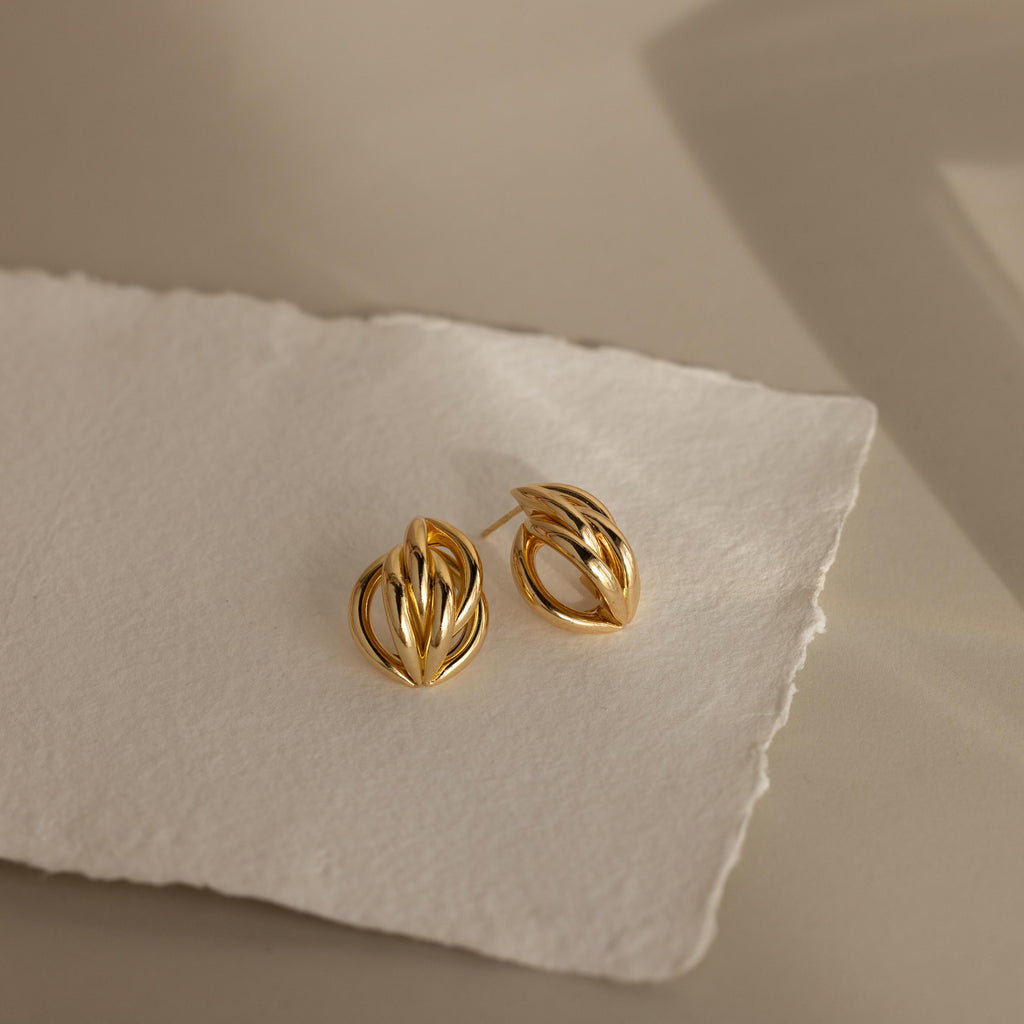 The Bold Multi Knot Studs feature an abstract, layered knot design and are displayed on textured white paper under soft lighting, highlighting their modern elegance.