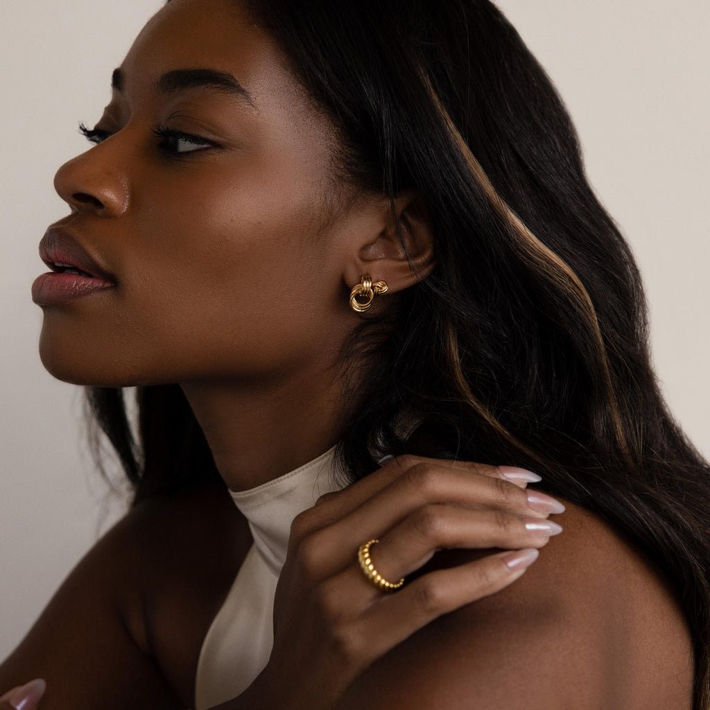 Woman with long dark hair exudes quiet luxury, wearing Micro Triple Knot Earrings and a ring as she poses in profile against a neutral background.