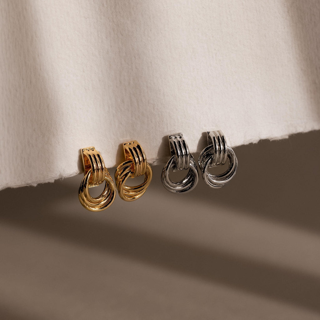 The Micro Triple Knot Earrings, in gold and silver pairs, exude quiet luxury. Shown on a textured white surface, these interlocking hoops echo the timeless elegance of spiral knot designs.