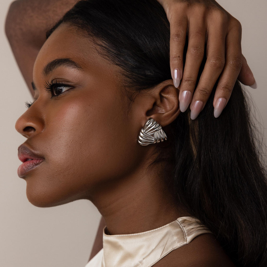 A woman with long hair wears Bold Triangle Knot Studs—shiny, sculpted silver earrings that catch the light as she touches her head, making a true statement accessory.