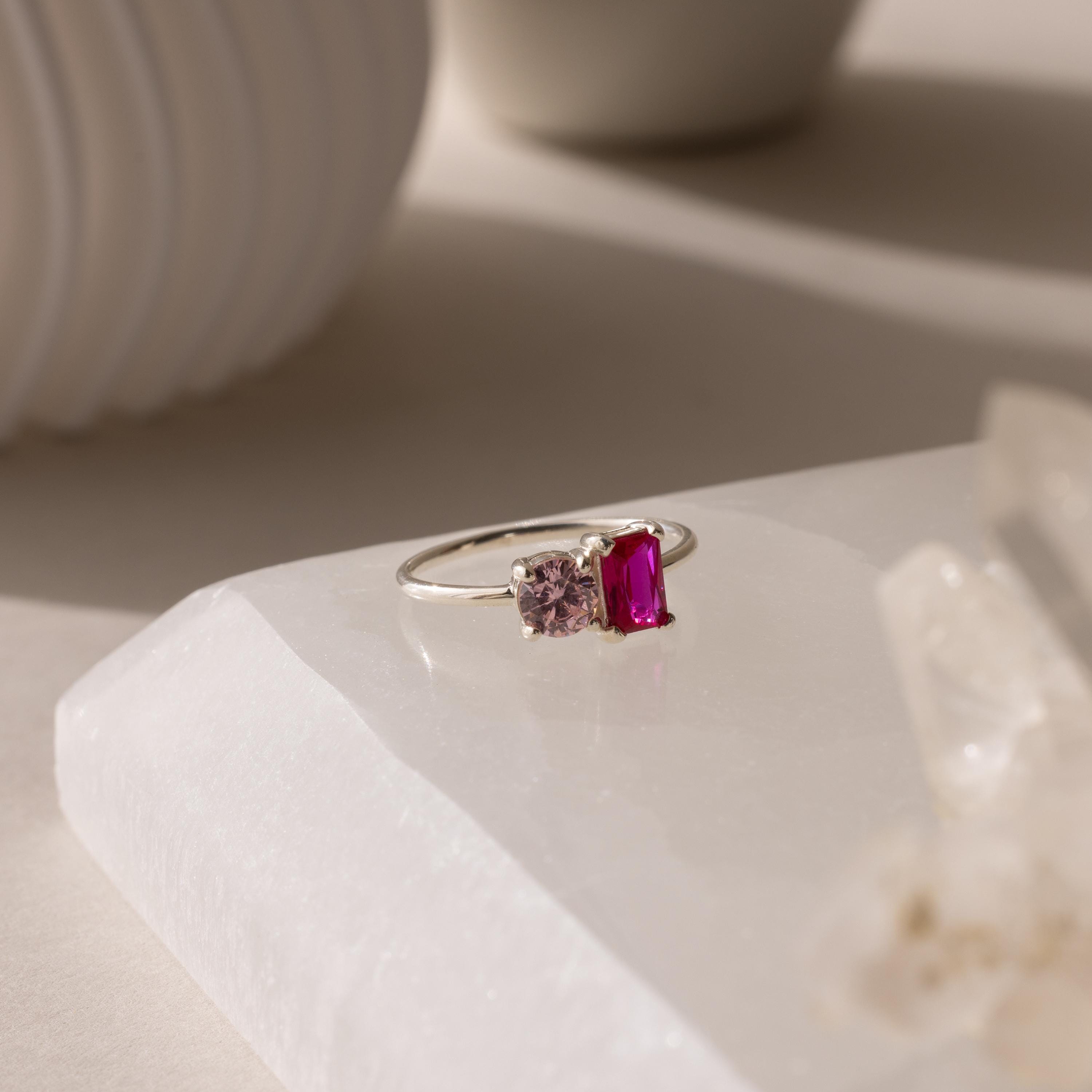 The Toi et Moi Emerald Birthstone Ring features a silver band with two rectangular gemstones—one pink, one red—displayed elegantly on a white stone surface.