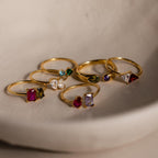 Six Open Toi et Moi Birthstone Rings with vibrant gemstones are displayed on a curved, light ceramic surface.