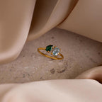 The Open Toi et Moi Birthstone Ring, featuring green and clear gems, is displayed on a stone surface with beige fabric elegantly draped around it.