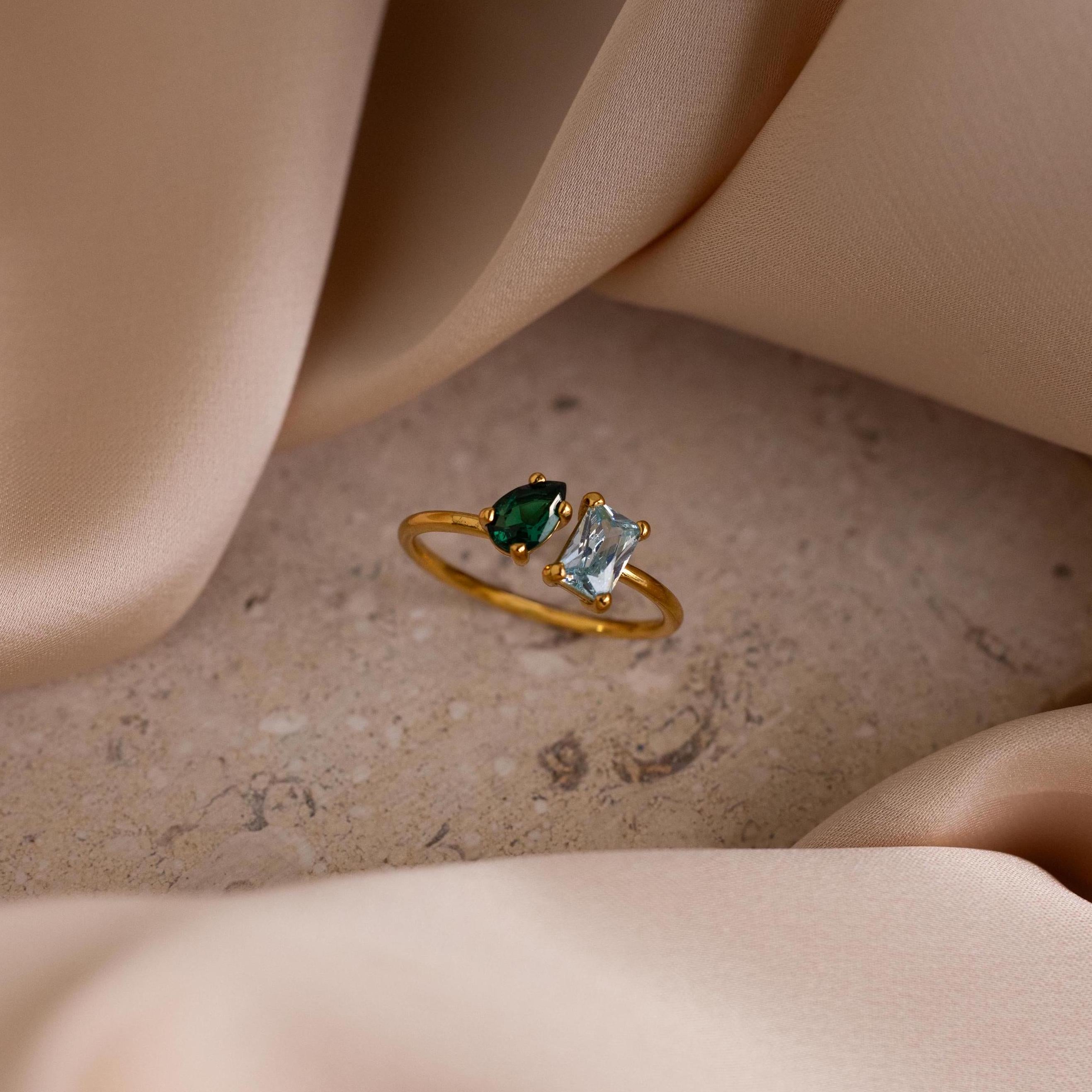 The Open Toi et Moi Birthstone Ring, featuring green and clear gems, is displayed on a stone surface with beige fabric elegantly draped around it.