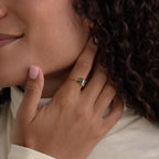 A person with curly hair touches their neck, wearing the Open Toi et Moi Birthstone Ring—a gold band set with green and white stones.