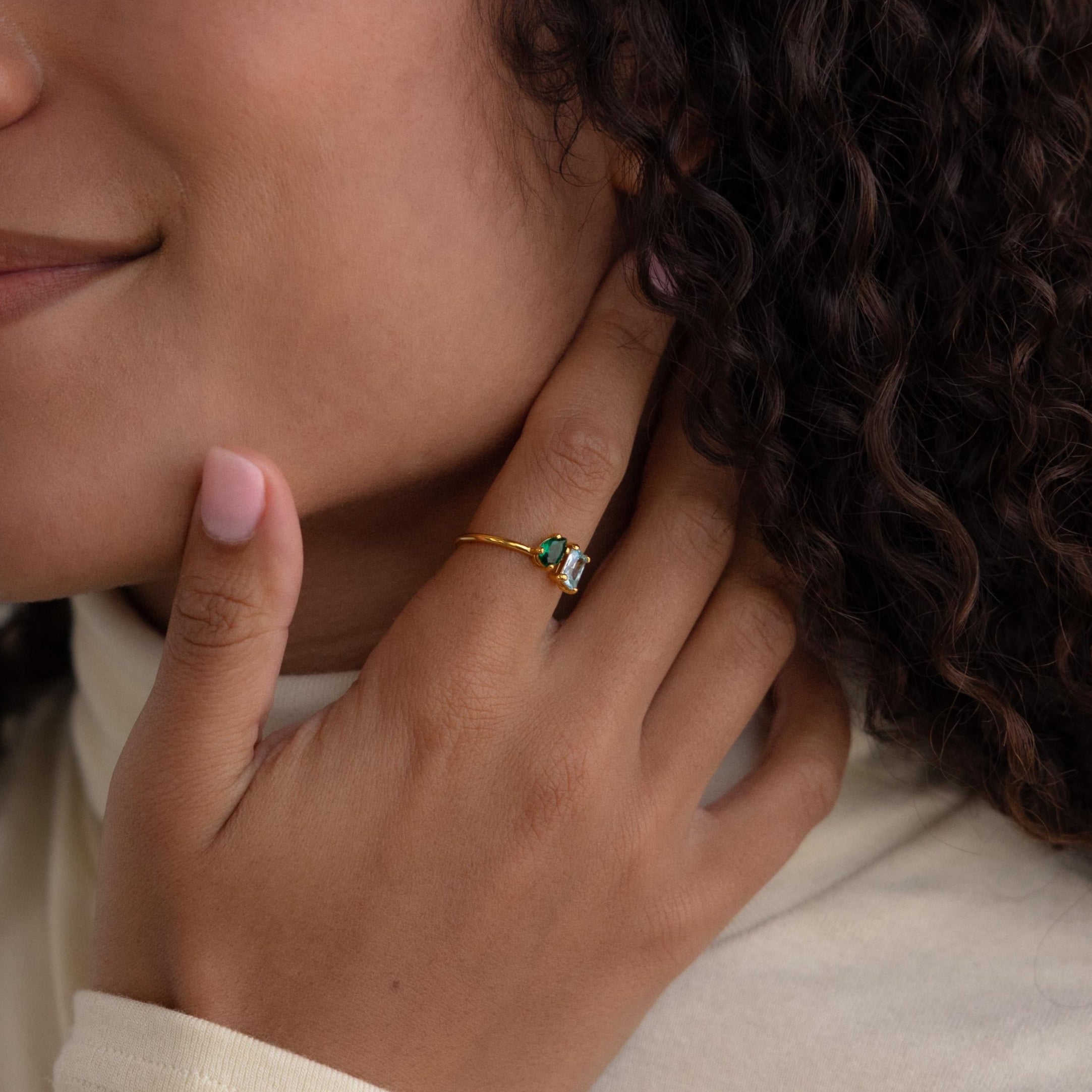 A person with curly hair touches their neck, wearing the Open Toi et Moi Birthstone Ring—a gold band set with green and white stones.