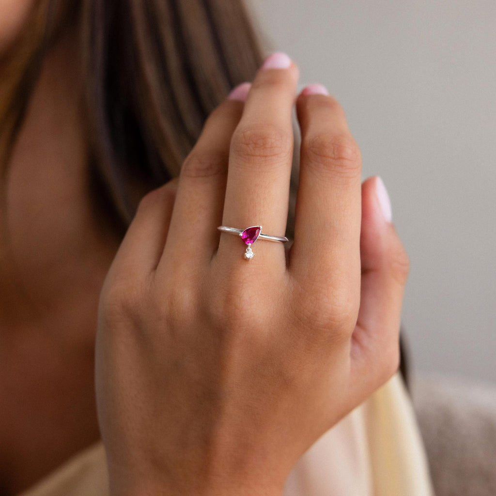 A hand wears the Duo Birthstone Drop Ring, a silver band featuring a red gemstone and a small round diamond charm on the ring finger.