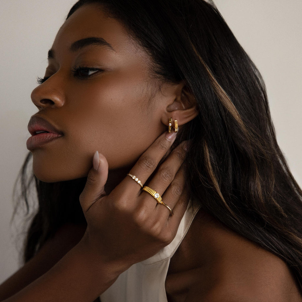 A woman with long hair wears the Diamond Ribbed Huggies—dainty hoops featuring a subtle ribbed design—adding an elegant touch as she gently touches her ear and chin, gazing to the side.