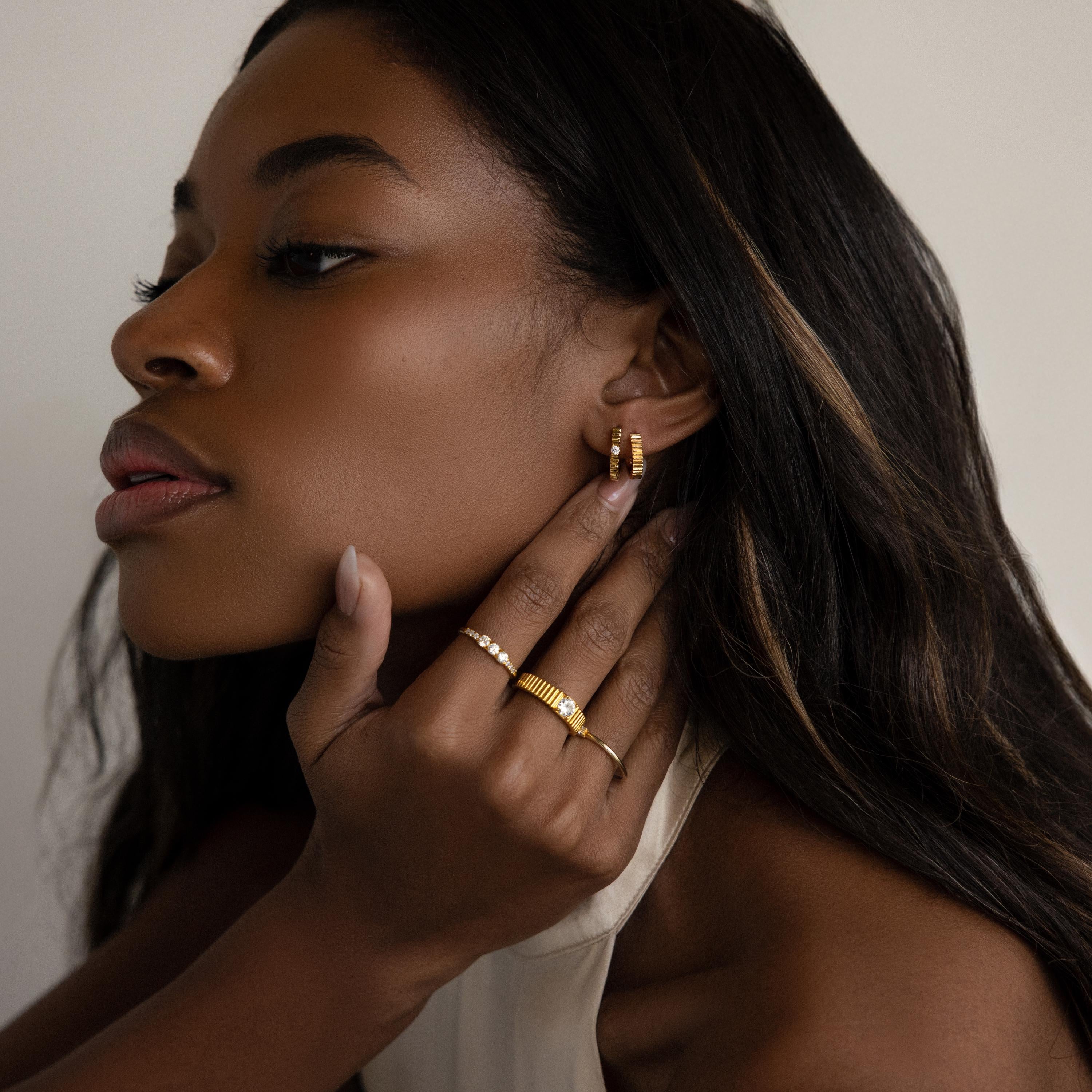 A woman with long hair wears the Diamond Ribbed Huggies—dainty hoops featuring a subtle ribbed design—adding an elegant touch as she gently touches her ear and chin, gazing to the side.