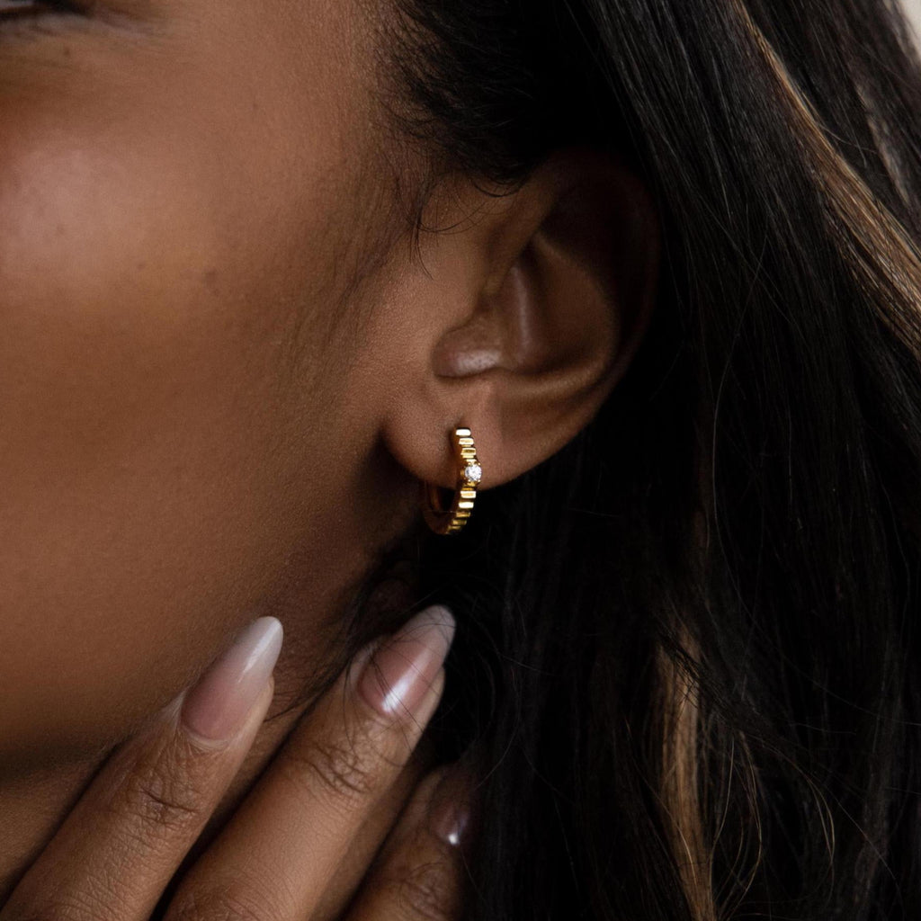 Close-up of a woman’s ear adorned with Diamond Ribbed Huggies; she gently touches her neck near her jaw, displaying the delicate ribbed design of the hoops.