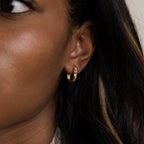 Close-up of a woman's ear adorned with Hammered Rectangle Hoops—small gold earrings with a textured finish—set against her long dark hair.