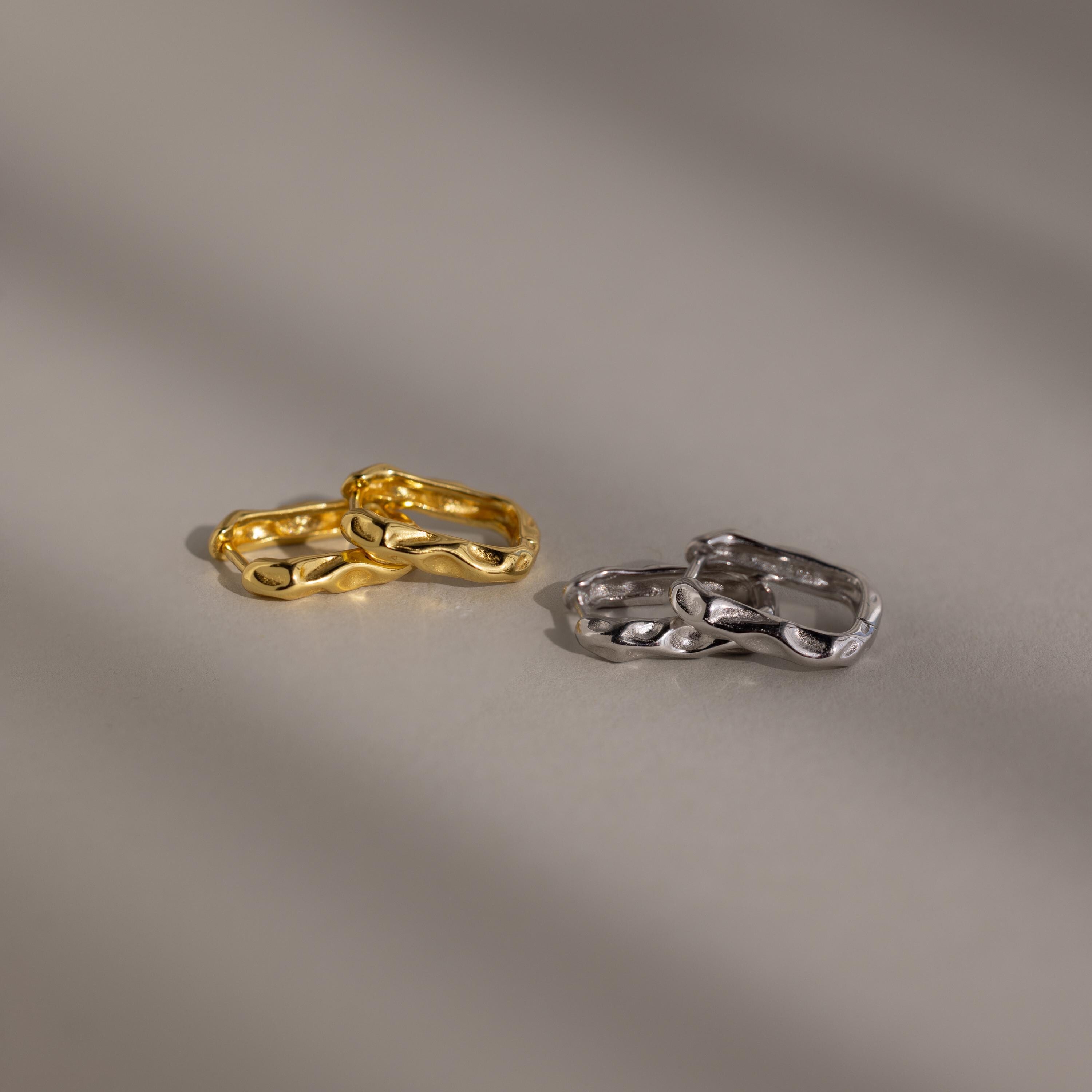 The Hammered Rectangle Hoops, featuring gold and silver textured geometric designs, are displayed side by side on a smooth, neutral surface.