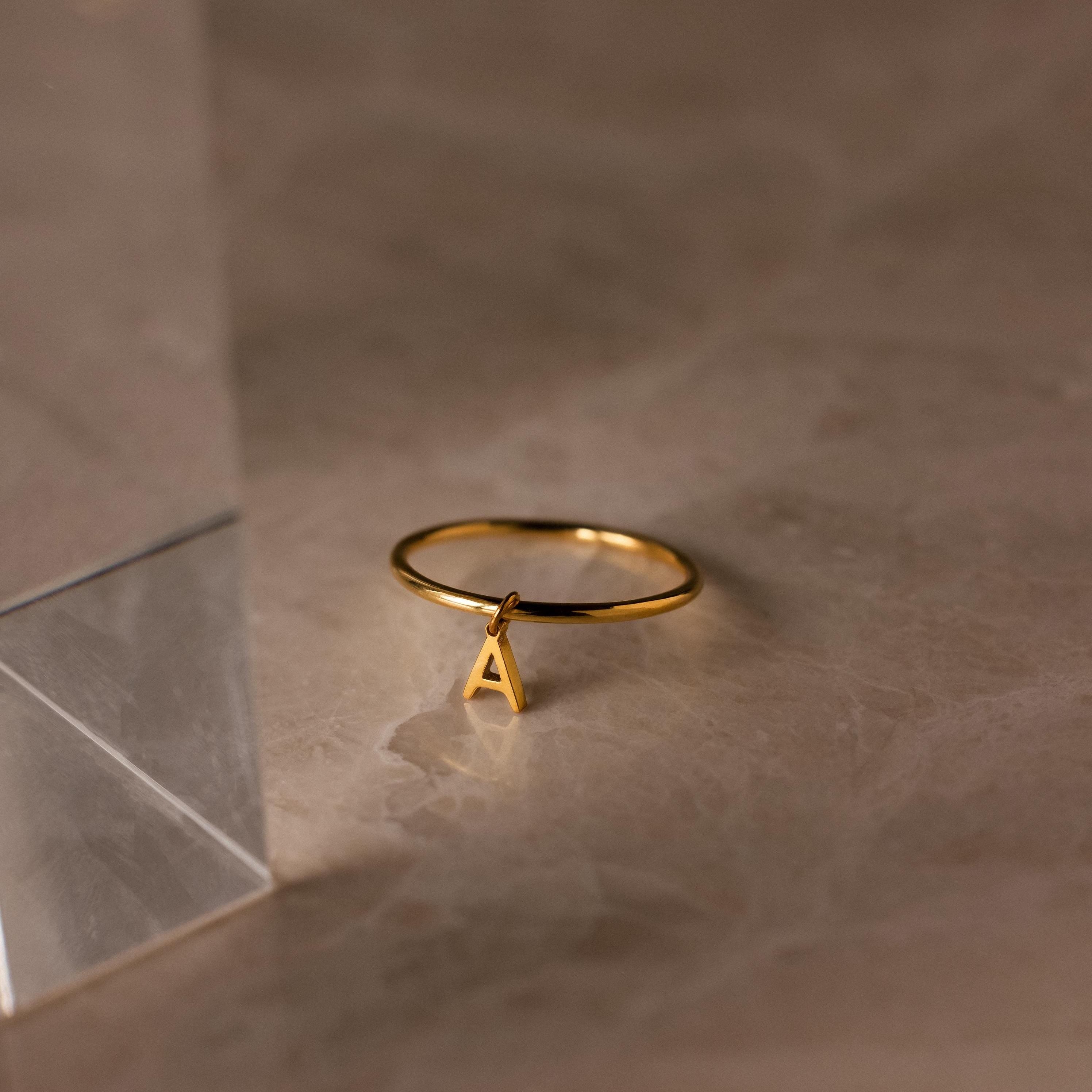 The Dainty Initial Drop Ring features a gold band with a dangling letter A charm, creating a personalized touch that shines beautifully on a marble surface.