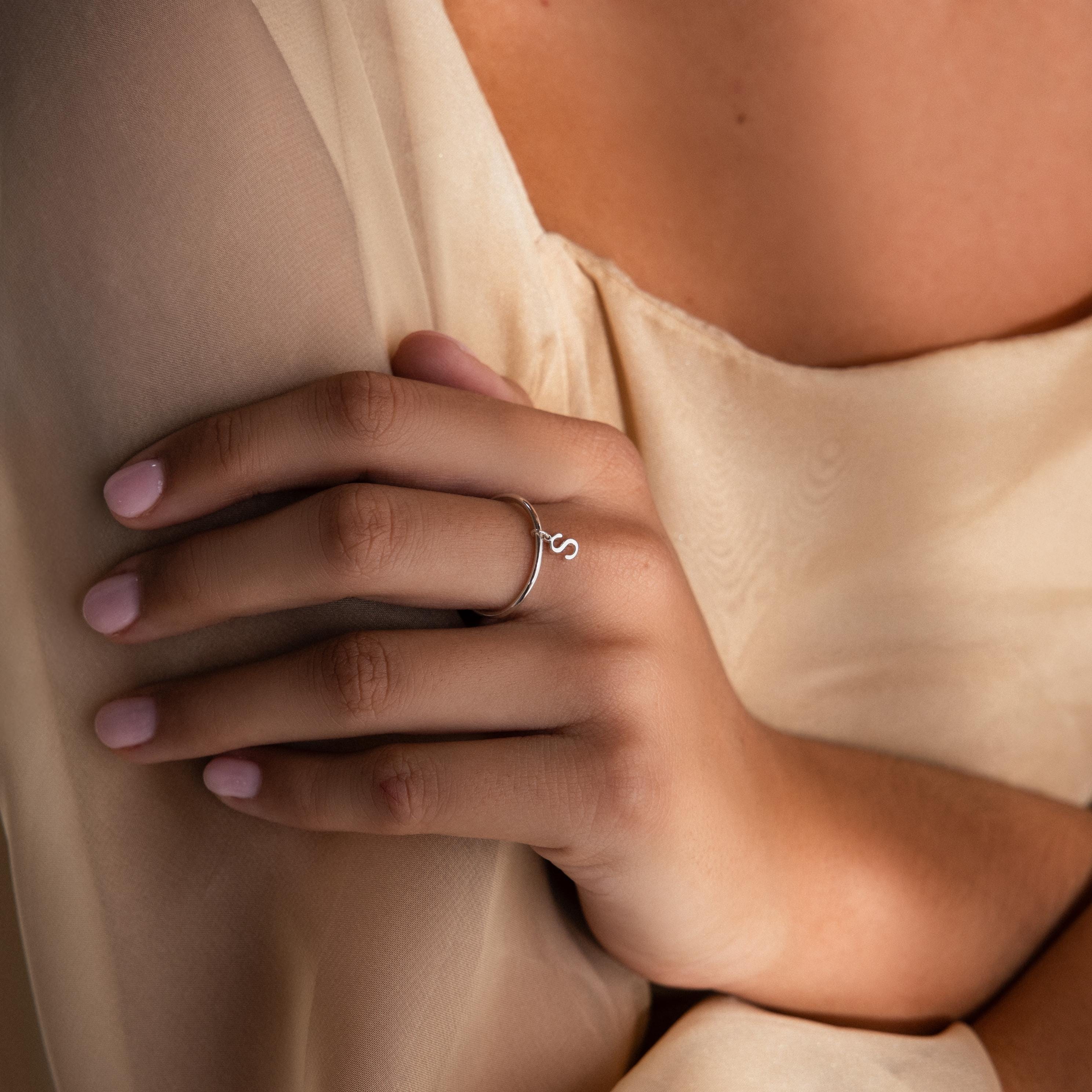 A hand with light pink nails wears the Dainty Initial Drop Ring, featuring an "S" initial, resting on beige fabric—a charming example of personalized jewelry.