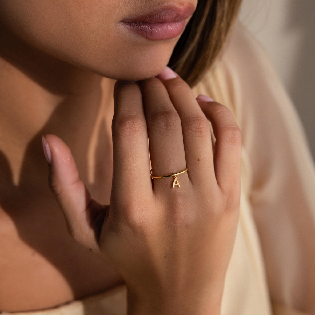 A woman touches her chin while wearing the Dainty Initial Drop Ring, which features a gold A charm for a personalized jewelry touch.