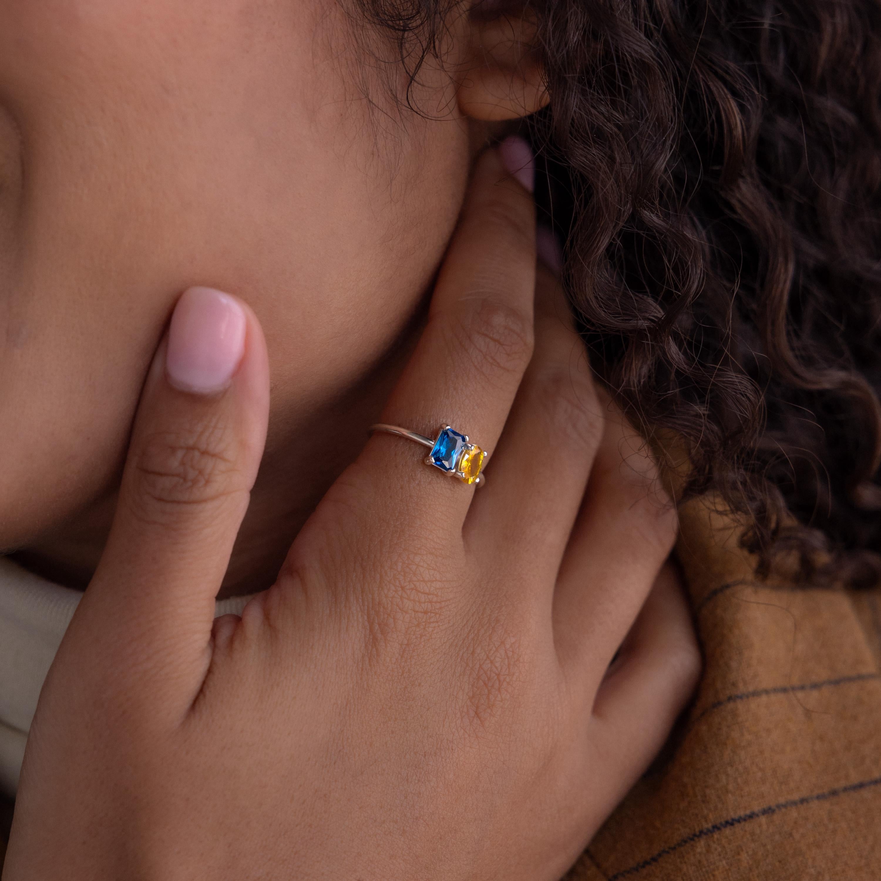 Close-up of a hand wearing the Toi et Moi Oval Birthstone Ring with blue and yellow stones, touching the side of the face.