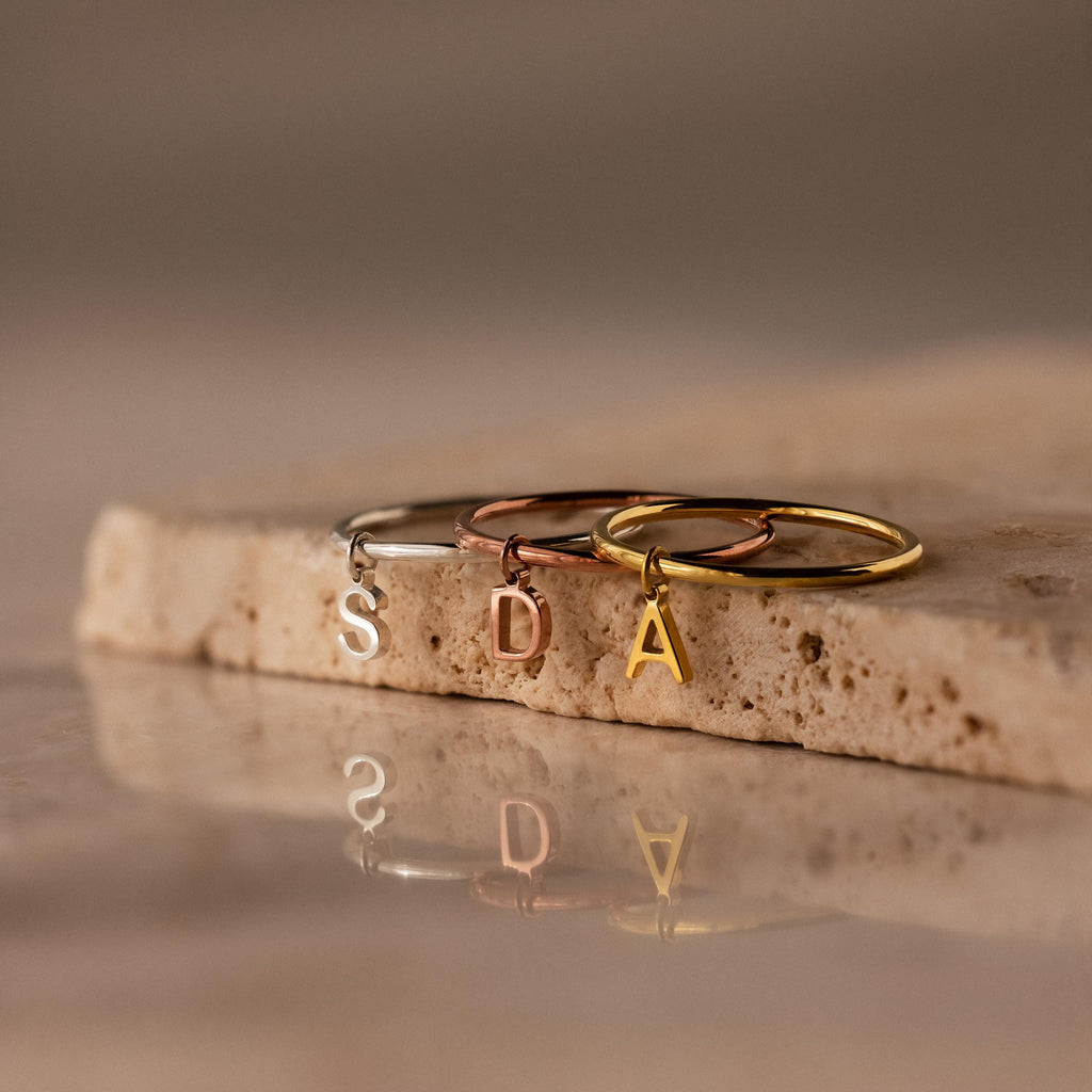 Three Dainty Initial Drop Rings in gold, silver, and rose gold with S, D, and A letter charms rest on a stone surface—perfect personalized jewelry for any occasion.