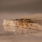 Three Dainty Initial Drop Rings in gold, silver, and rose gold with S, D, and A letter charms rest on a stone surface—perfect personalized jewelry for any occasion.