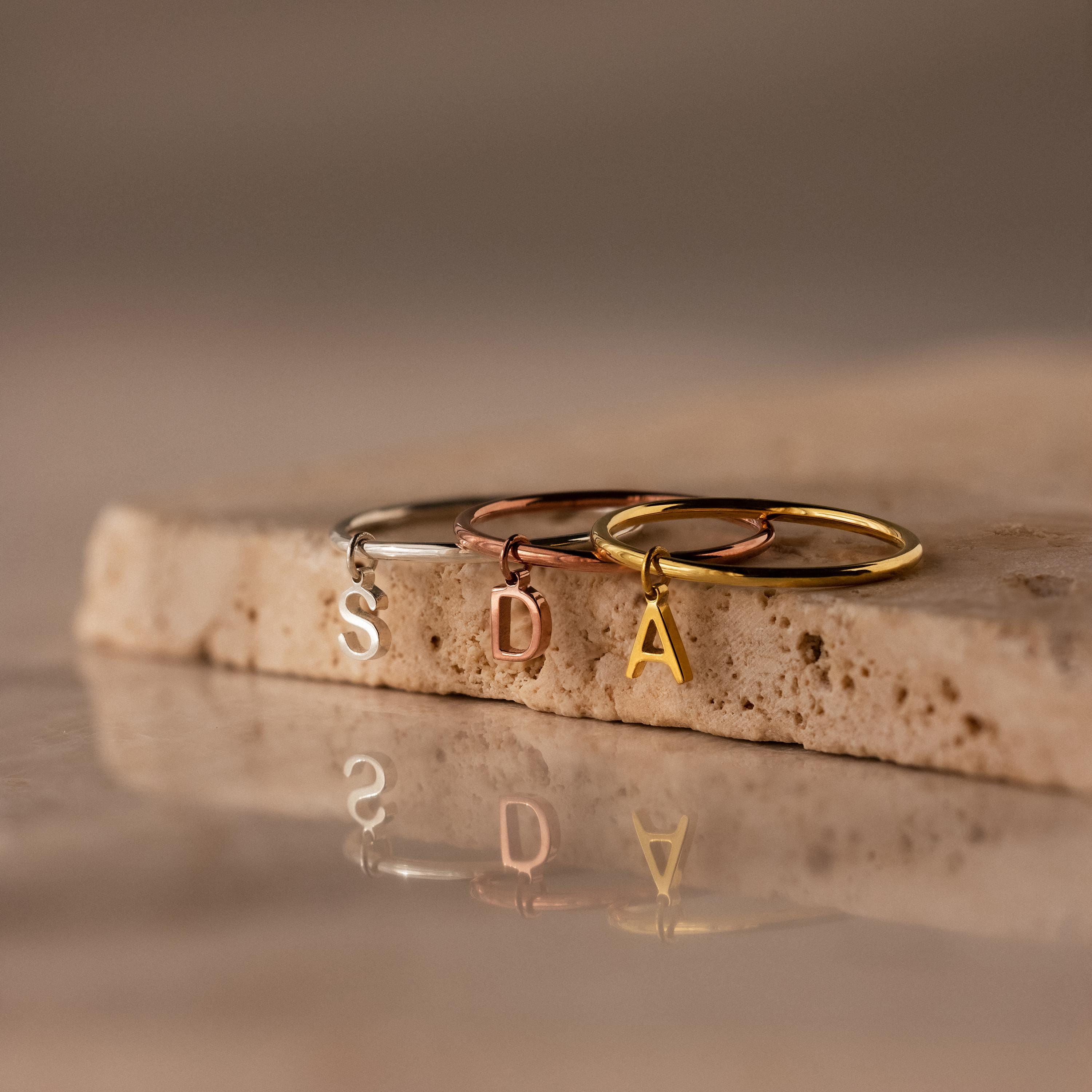Three Dainty Initial Drop Rings in gold, silver, and rose gold with S, D, and A letter charms rest on a stone surface—perfect personalized jewelry for any occasion.
