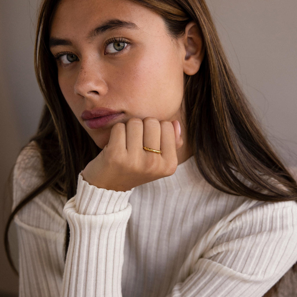 Woman with long brown hair wearing a white ribbed top and a gold Dainty Morse Code Ring, resting her chin on her hand.