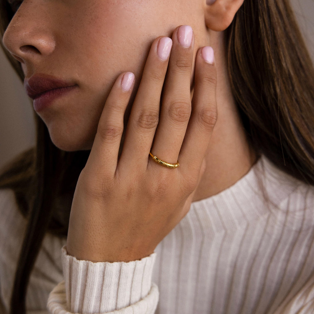 A woman wears a Dainty Morse Code Ring, touching her face with manicured fingers while dressed in a cream ribbed sweater.