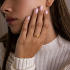 A woman wears a Dainty Morse Code Ring, touching her face with manicured fingers while dressed in a cream ribbed sweater.