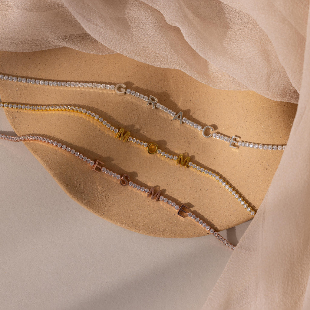 A Mama Tennis Letter Bracelet with gold letters spelling MAMA rests on a beige fabric background—an ideal example of customized jewelry, perfect for stylish gift giving.