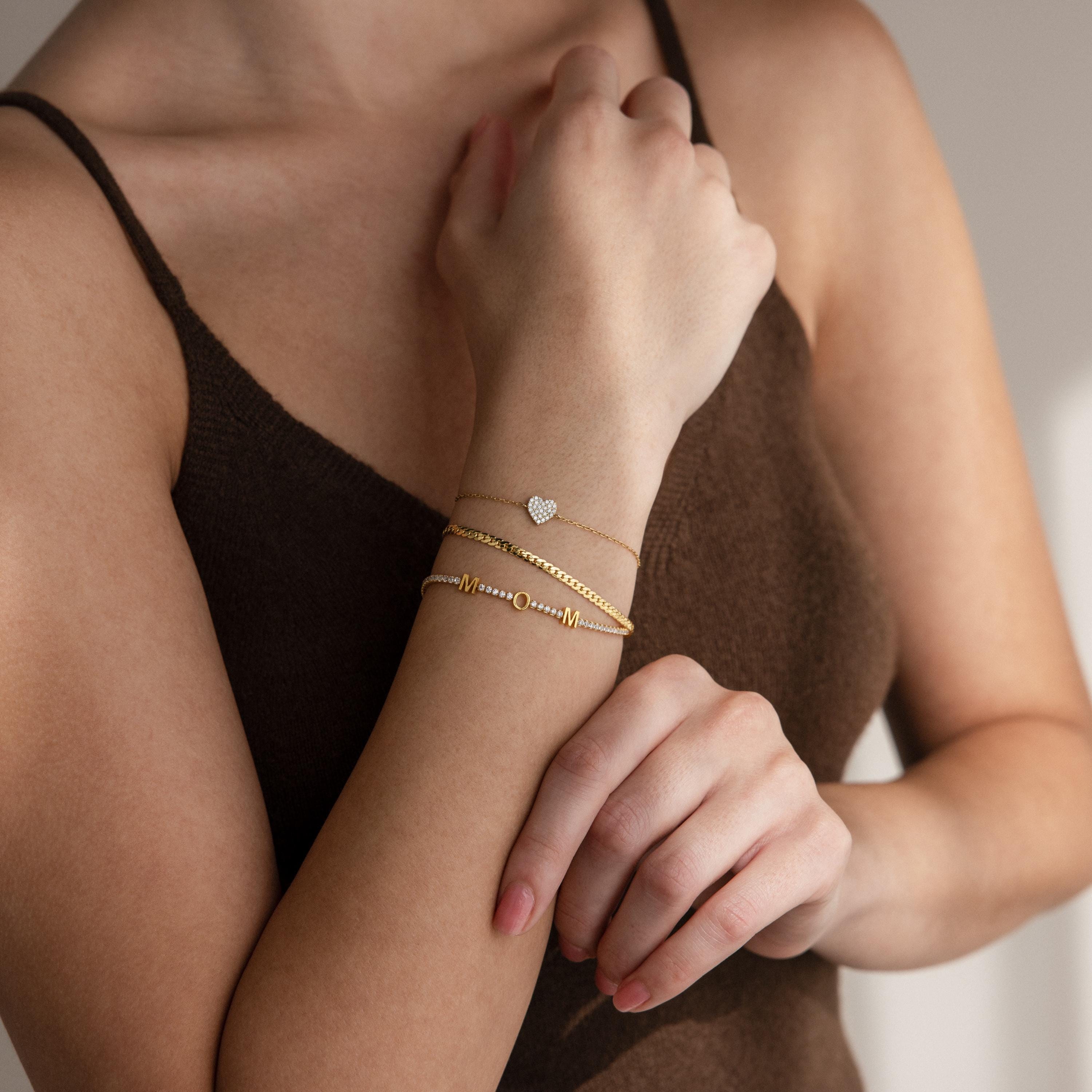A woman in a brown top wears three gold bracelets, including the Mama Tennis Letter Bracelet with a heart charm—an ideal choice for fans of personalized jewelry and thoughtful gift giving.