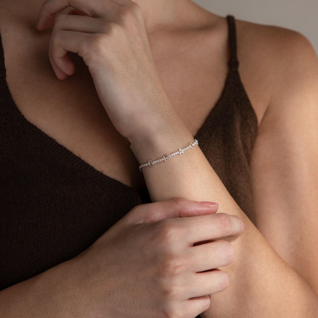 Someone in a brown tank top wears the Mama Tennis Letter Bracelet, delicately touching their wrist—ideal for fans of personalized jewelry and meaningful gift-giving.