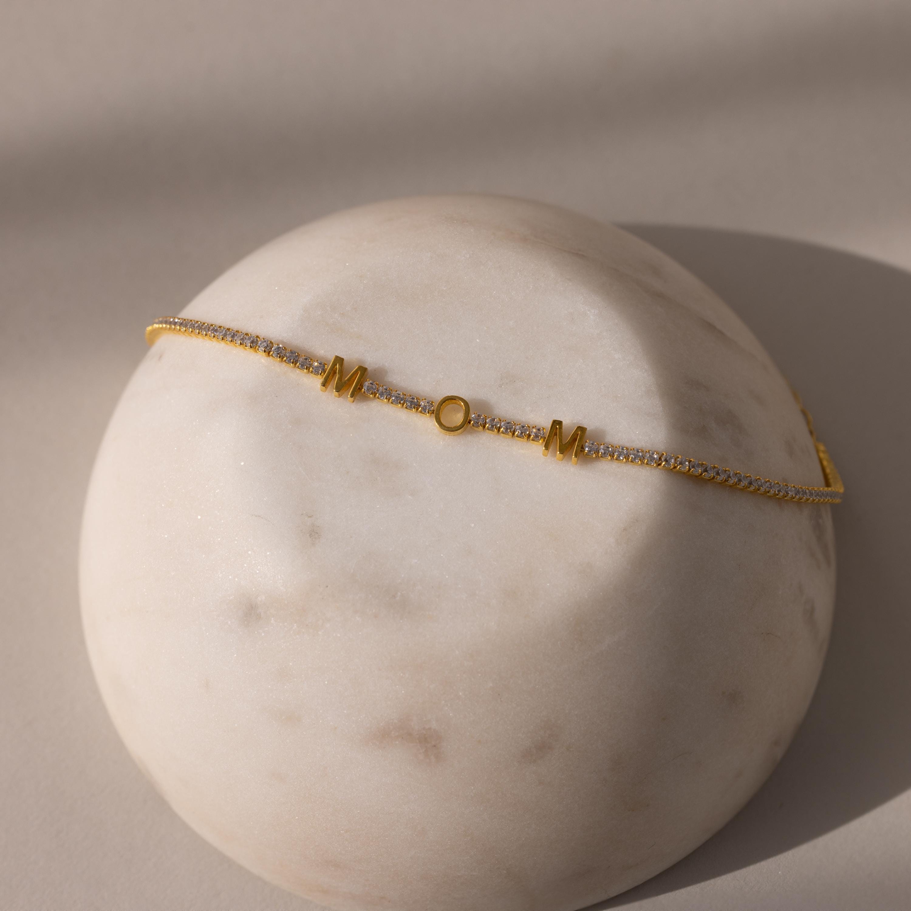 The Mama Tennis Letter Bracelet features "MOM" and a circle charm on delicate gold, displayed on a round marble-like stone—an ideal customized jewelry piece for thoughtful gifting.