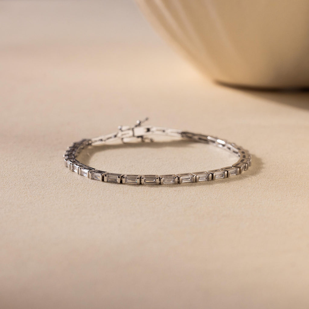 An elegant jewelry piece, the Claudia Baguette Tennis Bracelet features rectangular silver links with baguette diamonds, displayed on a beige surface against a softly blurred background.