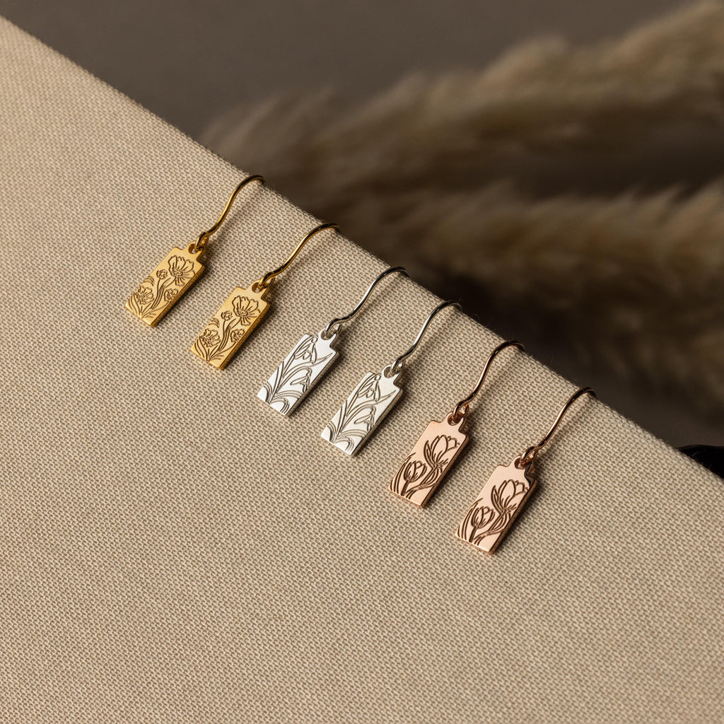 The Rectangle Birth Flower Earrings are available in gold, silver, and rose gold with floral engravings, beautifully displayed on beige fabric for a personalized jewelry touch.