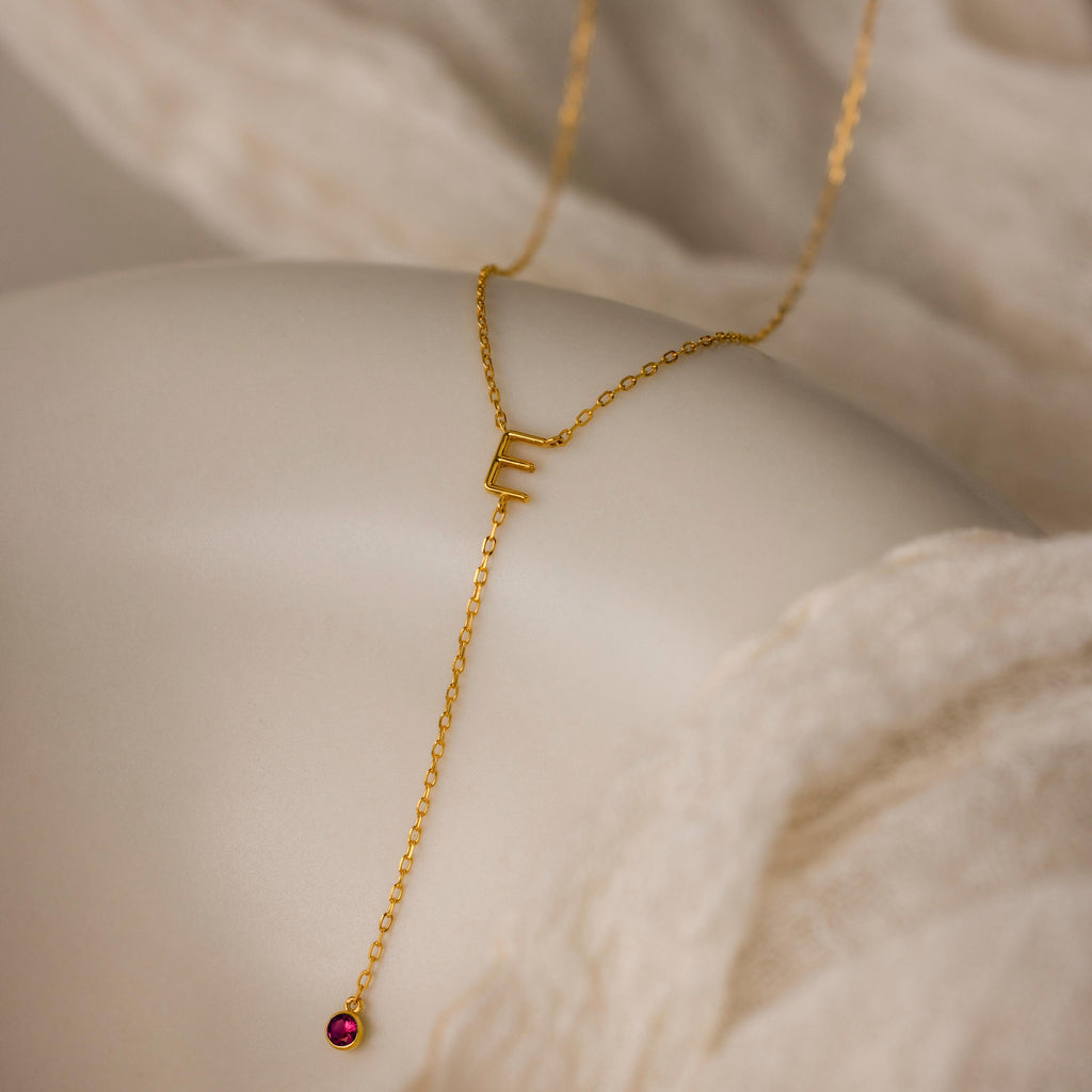 The Initial Birthstone Lariat Necklace, featuring an E charm and a small red gemstone on a white draped fabric, is a stunning example of personalized jewelry.