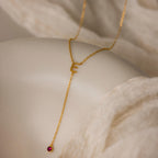 The Initial Birthstone Lariat Necklace, featuring an E charm and a small red gemstone on a white draped fabric, is a stunning example of personalized jewelry.