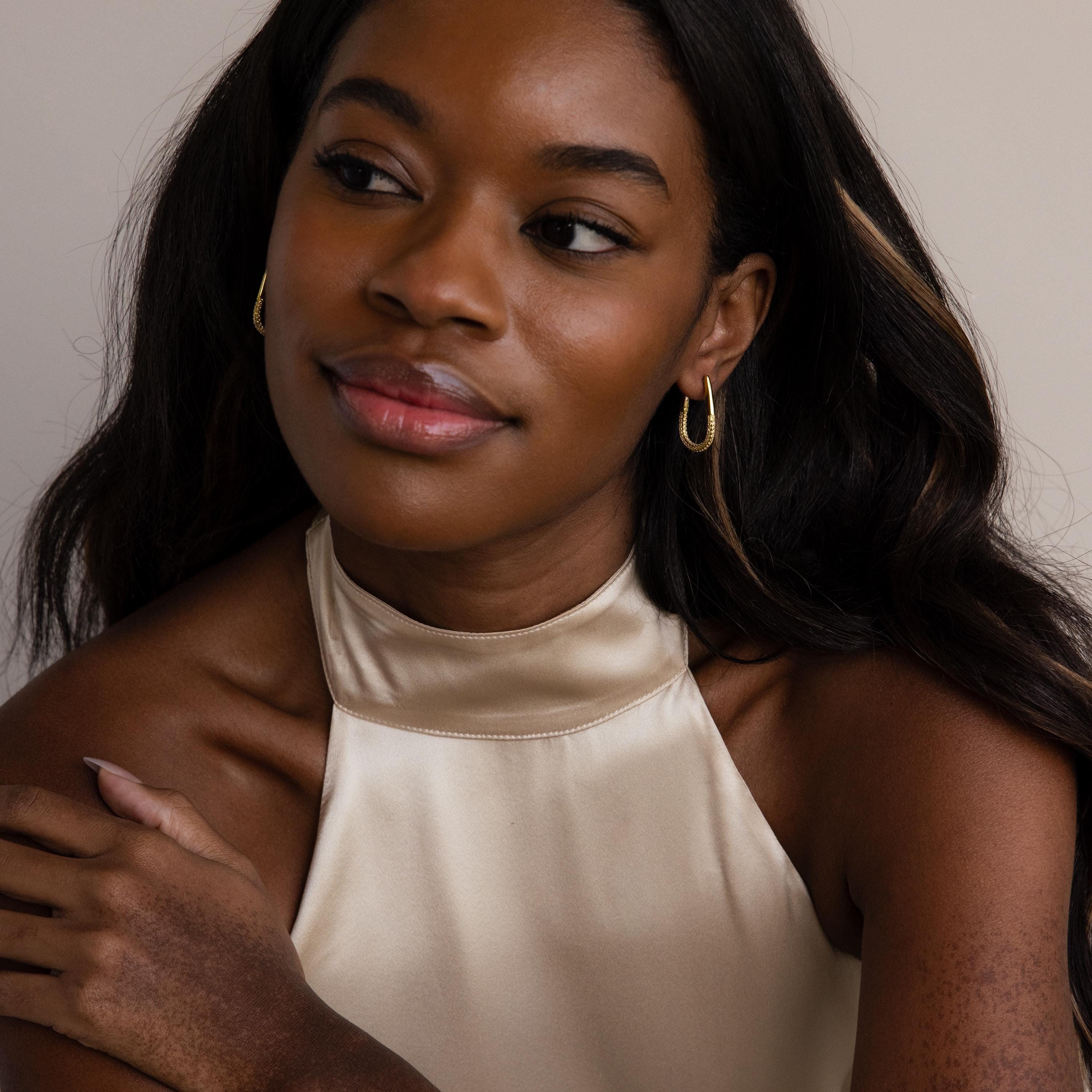 A woman with long dark hair, smiling softly, wears the Bold Pave Oval Hoops and a cream satin halter top.