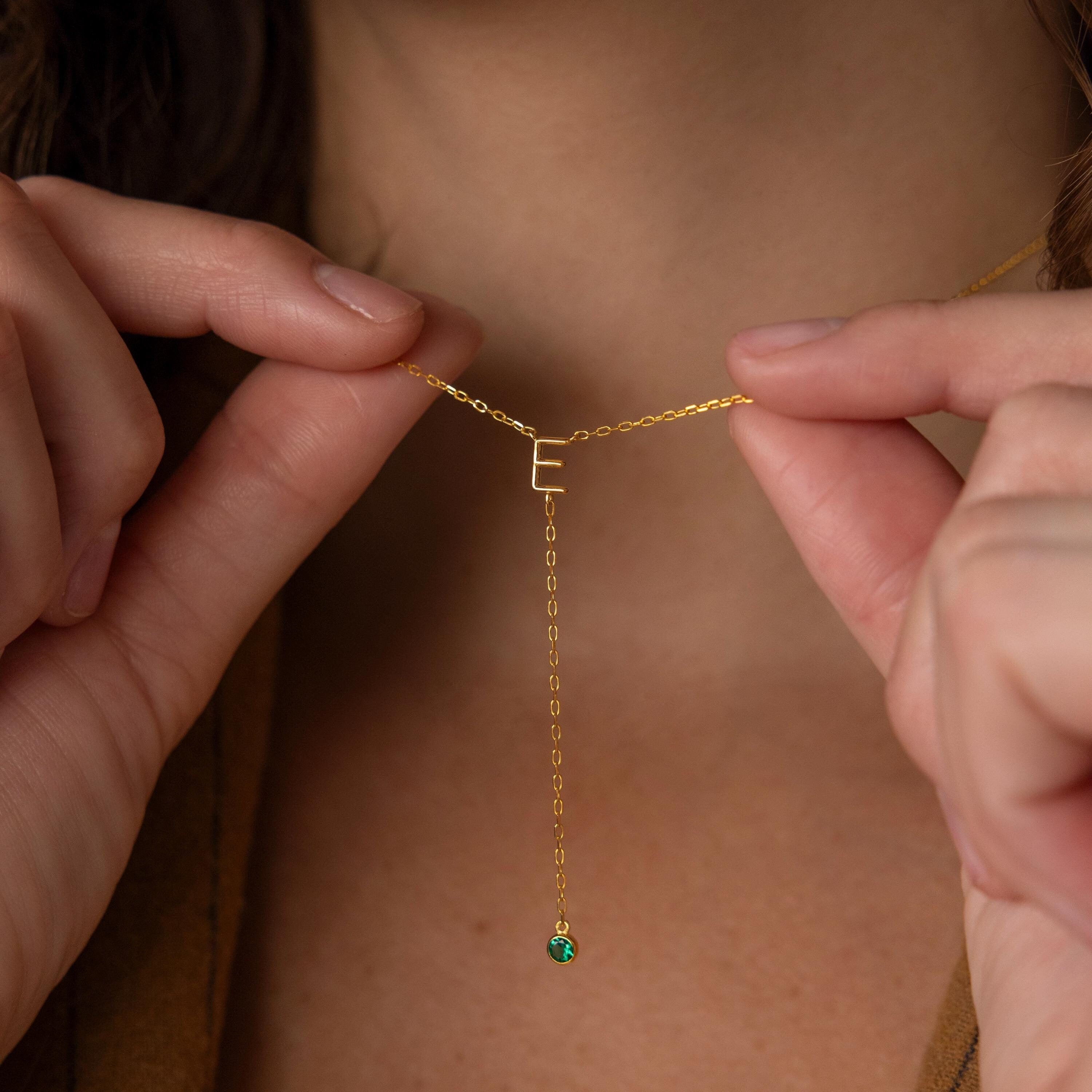 The Initial Birthstone Lariat Necklace features an "E" initial and a dangling green gemstone, showcased as a personalized accessory gently held near the wearer's neck.