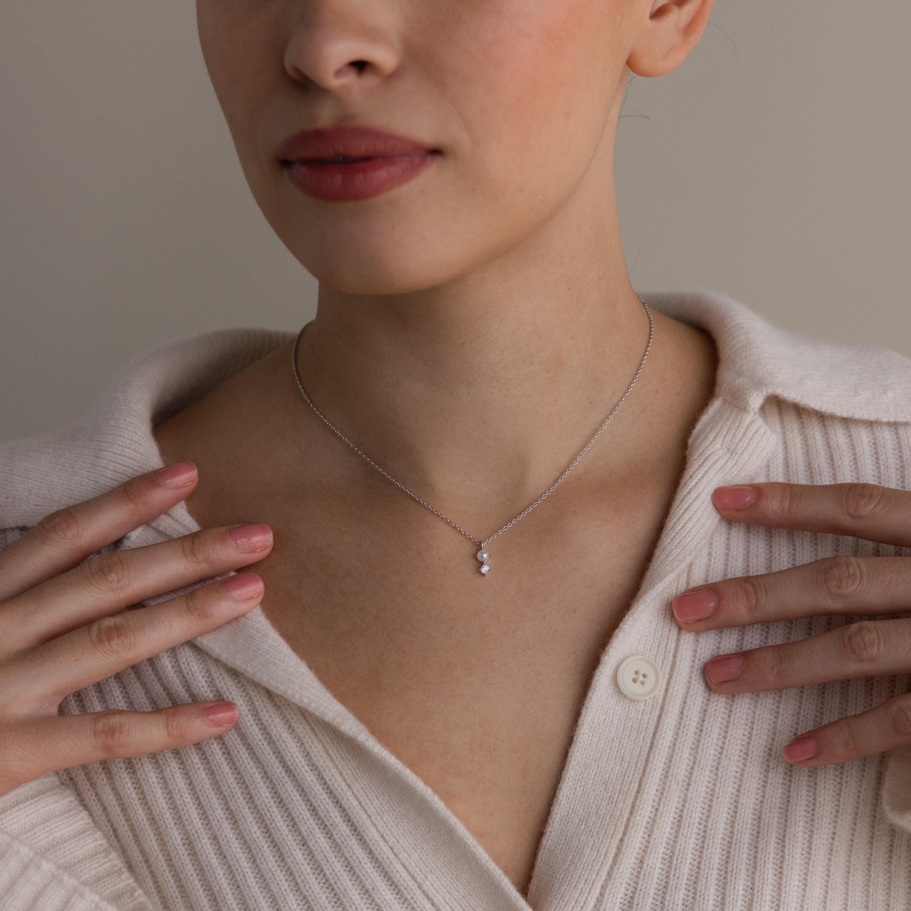 Woman wearing a cream sweater and a Pearl Diamond Drop Necklace, delicately touching the collar with both hands.