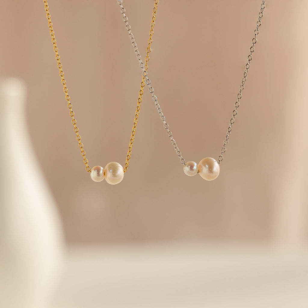 The Dainty Duo Pearl Necklace features two delicate pearls on gold and silver chains, displayed on beige—an elegant piece of jewelry and a thoughtful wedding gift.