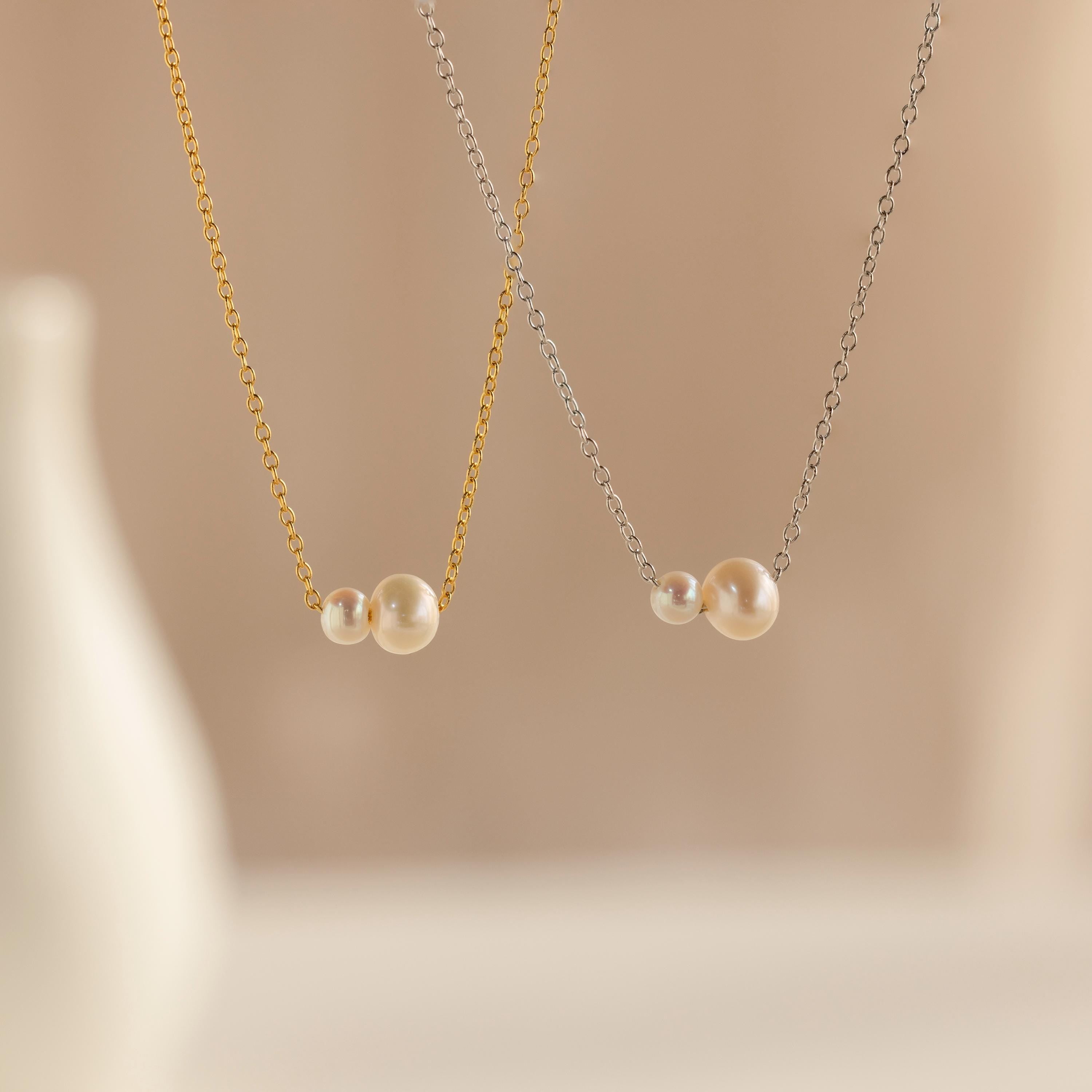 The Dainty Duo Pearl Necklace features two delicate pearls on gold and silver chains, displayed on beige—an elegant piece of jewelry and a thoughtful wedding gift.