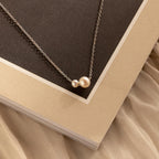 The Dainty Duo Pearl Necklace, featuring two gold pearls, rests on a textured beige and brown book cover with a satin backdrop—a perfect pick for dainty jewelry lovers or as a thoughtful wedding gift.