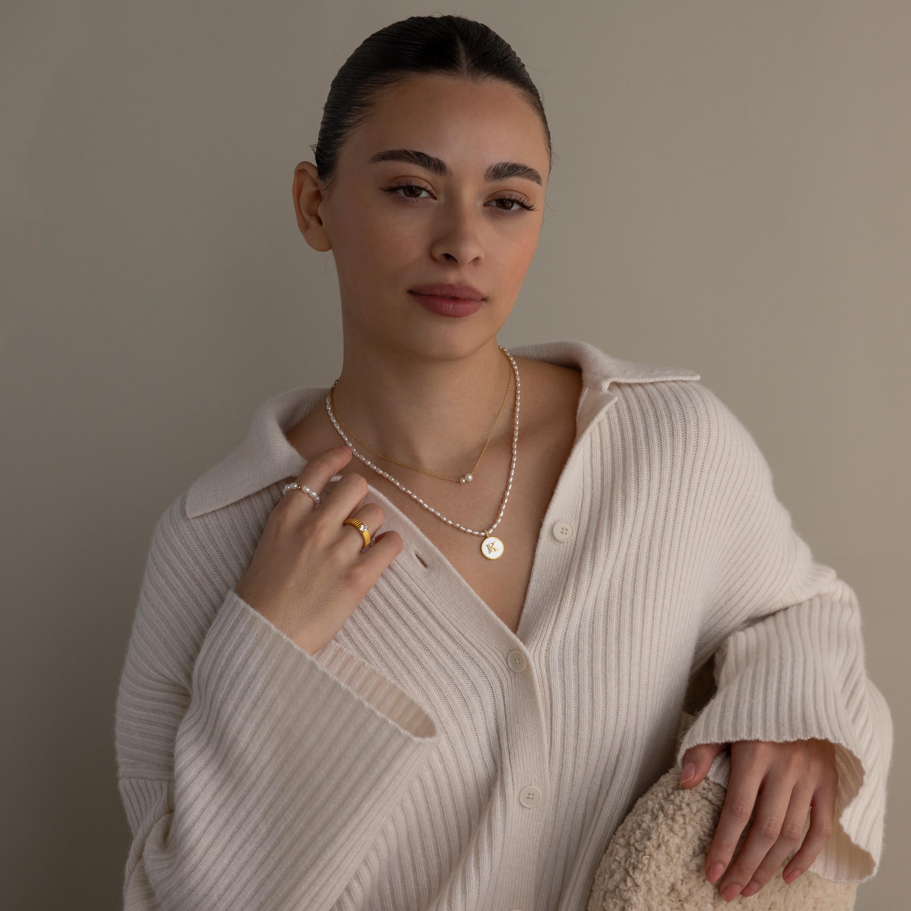Woman in a cream ribbed sweater poses against a neutral background, wearing dainty jewelry including the Dainty Duo Pearl Necklace and stylish rings.