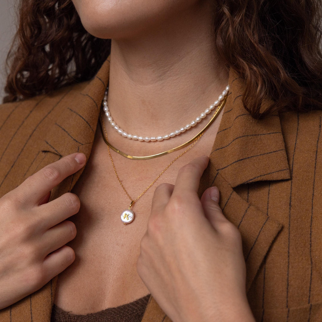 A woman wearing layered necklaces, including the Pearl Initial Pendant Necklace, and a brown pinstripe blazer, touches the gold pendant on her neck.