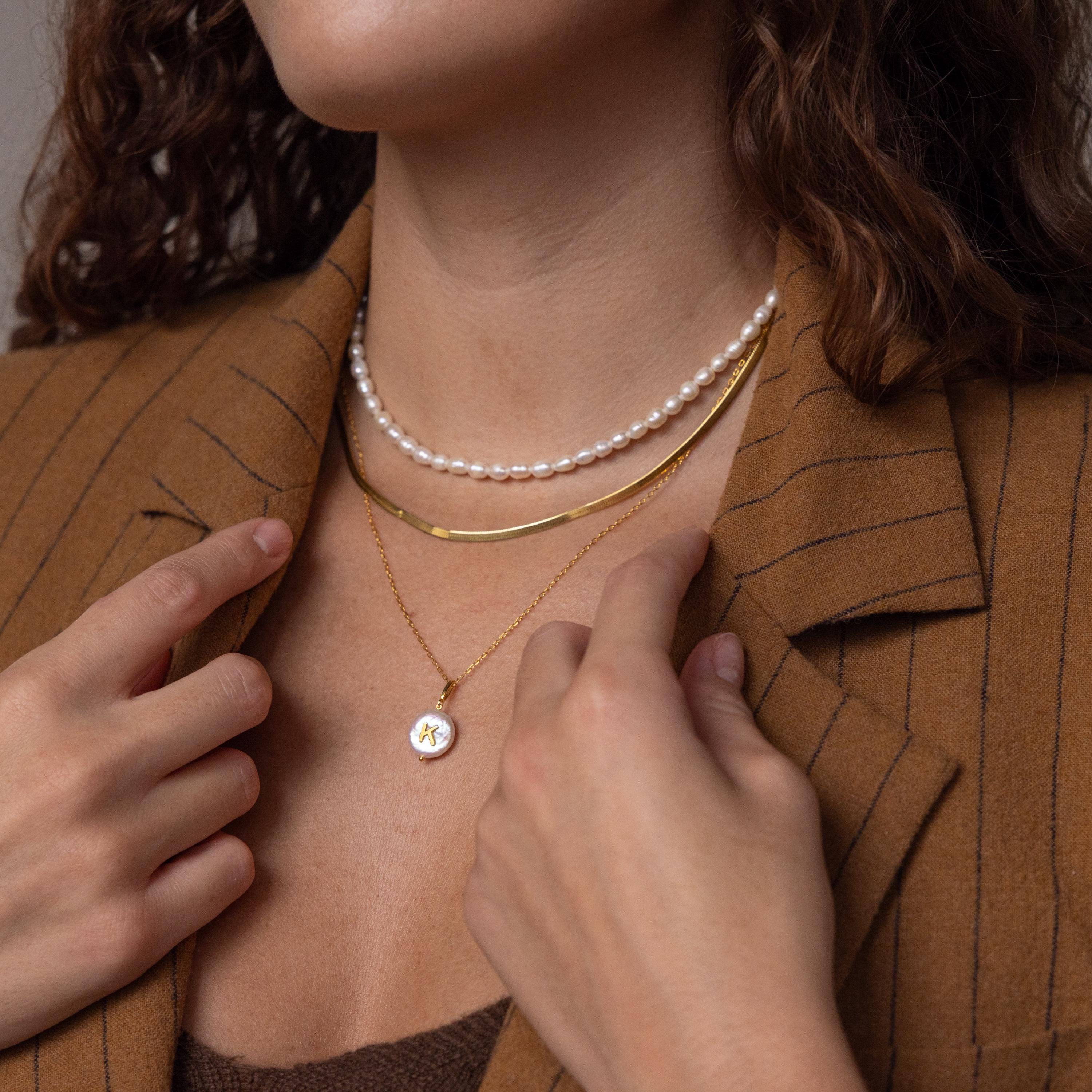 A woman wearing layered necklaces, including the Pearl Initial Pendant Necklace, and a brown pinstripe blazer, touches the gold pendant on her neck.