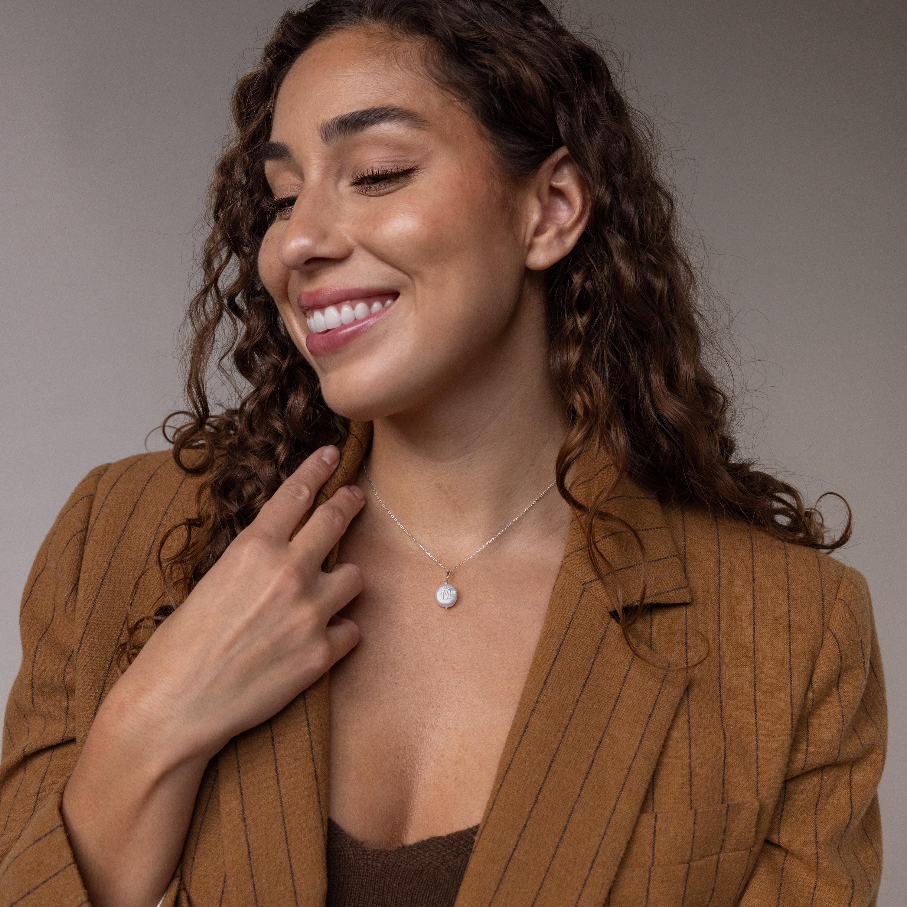 A smiling woman with curly hair, wearing a pinstripe blazer and the Pearl Initial Pendant Necklace, looks to the side.