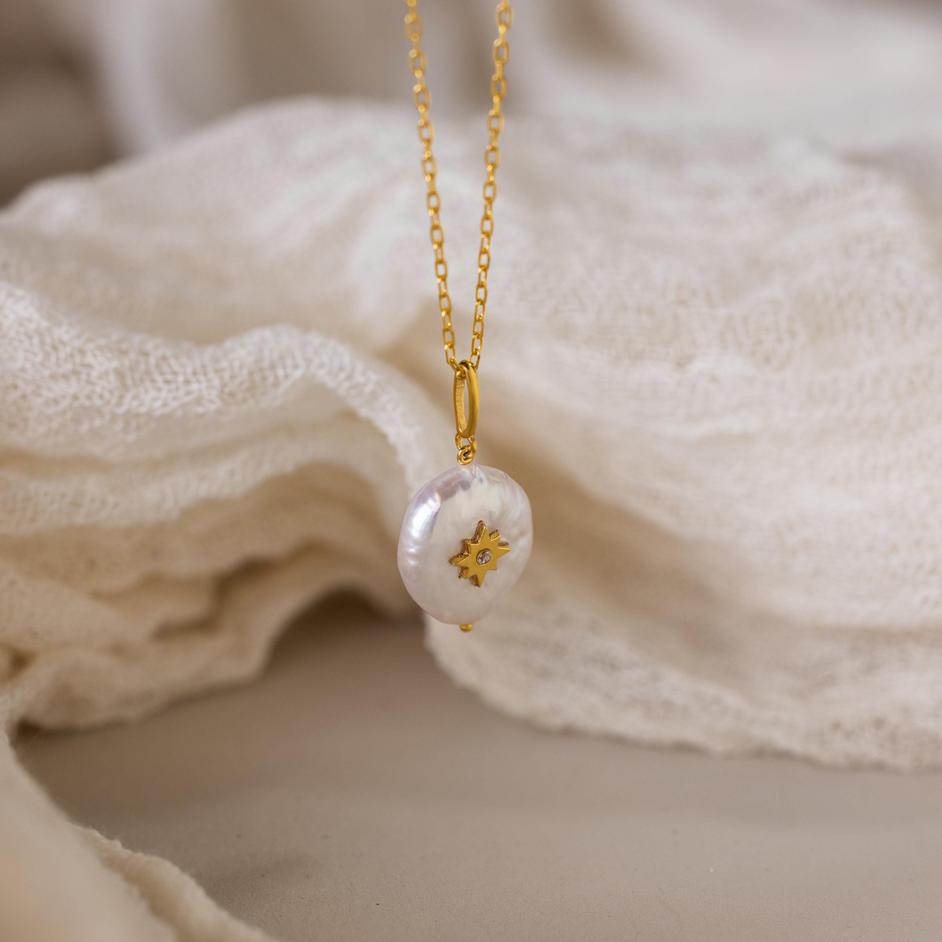 The Pearl Initial Pendant Necklace features a gold chain, a round freshwater pearl pendant, and a small gold star, displayed on soft beige fabric. Ideal for adding personalized elegance to any outfit.